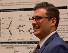 Dom, with glasses, short brown hair and brown mustache, smiling in quarter profile facing left and wearing a blue suit, standing in front of a research poster.